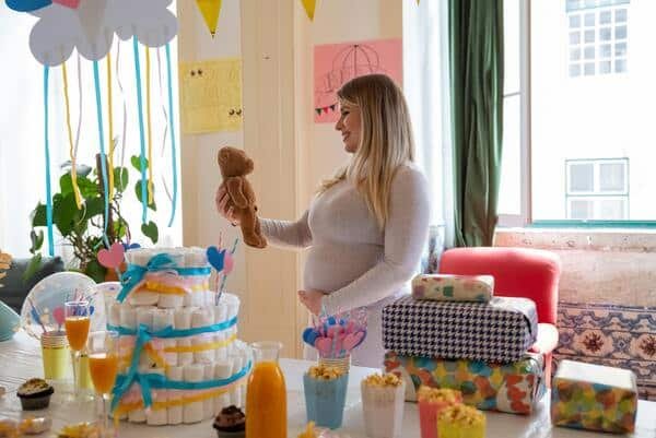 Madre embarazada sostiene en su mano un peluche mientras que con la otra mano acaricia su vientre. Enfrente de ella una mesa decorada para baby shower con postres, bebidas, regalos y globos.