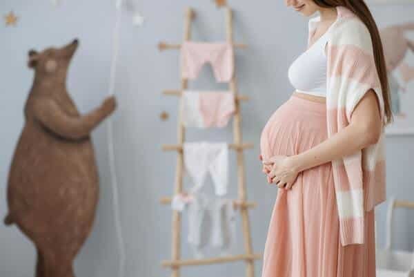 Mujer embarazada acariciando su vientre. Esta de pie en una habitación decorada con ropita para niña.