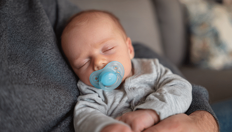 Bebé recién nacido en los brazos de papá, esta dormido y porta un chupón de color azul en su boca.