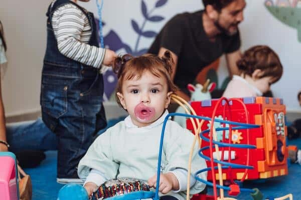 Niña con dos coletas jugando en un salón recreativo. La niña sostiene en su boca un chupón de color rosa.