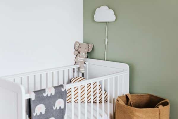 Cuna de bebe en una esquina de un cuarto Dentro de la cuna una almohada, cobija y un muñeco de elefante arriba de la cuna. Arriba de ella un lampara en forma de nube blanca.