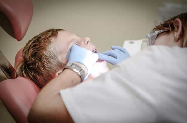 Niño reposa en silla de un dentista, mientras el dentista revisa su boca abierta.