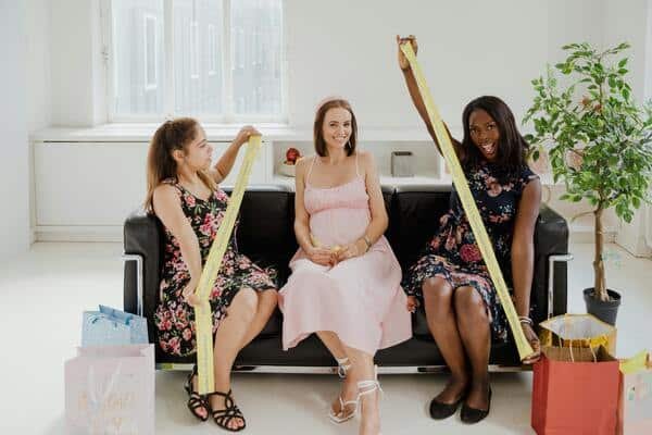 Mujer embarazada sentada en medio de un sofá, porta un vestido rosa y tacones blancos. Otras dos mujeres sentadas a su lado, ambas sostienen en sus manos una cinta de color amarillo. Enfrente de ellas regalos en el piso.