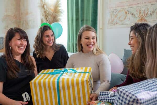 Cinco mujeres sentada en un sofá, la mujer de en medio carga un regalo gran entre sus brazos. 