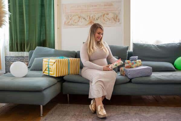 Mujer joven embarazada, sentada en el sofá de una casa moderna. Esta observando algunos regalos de su baby shower que están acomodados en el sofá.