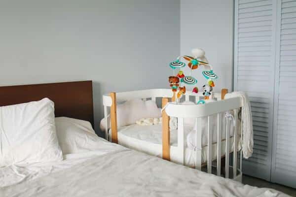 Habitación con cama matrimonial, a su lado una cuna de bebé. La cuna esta decorada con almohadillas a los costados en forma de luna, sabanas blancas y muñecos colgantes. La cama matrimonial también acomodada con sabanas blancas.